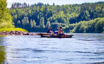 Två män sitter ombord på en mindre timmerflotte utan tältkonstruktion. En varm solig sommardag. Flotte glider lugnt och fint nedströms, männen doppar fötterna i vattnet. Mycket grönska längs strandkanten.