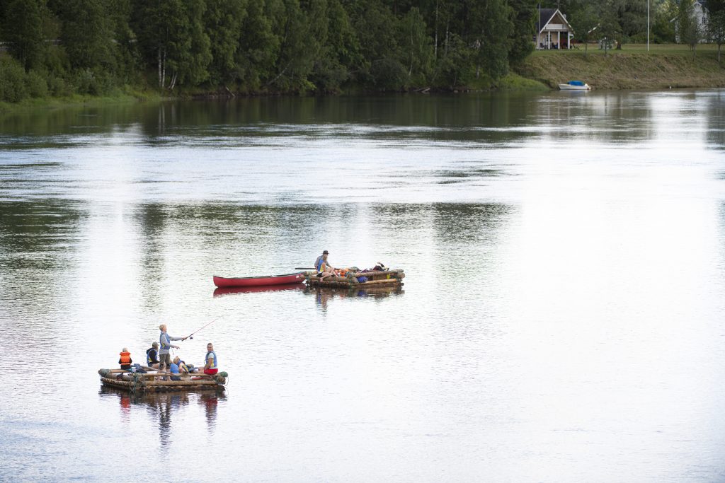Två mindre flottar utan tältkonstruktion flyter med lite avstånd till varandra på Klarälven. Vattnet blänkar vitt av solen. På ena flotten sitter och står flera personer, en pojke försöker fiska. Andra flotten har med en kanot som jolle. I bakgrunden står ett hus nära älven.