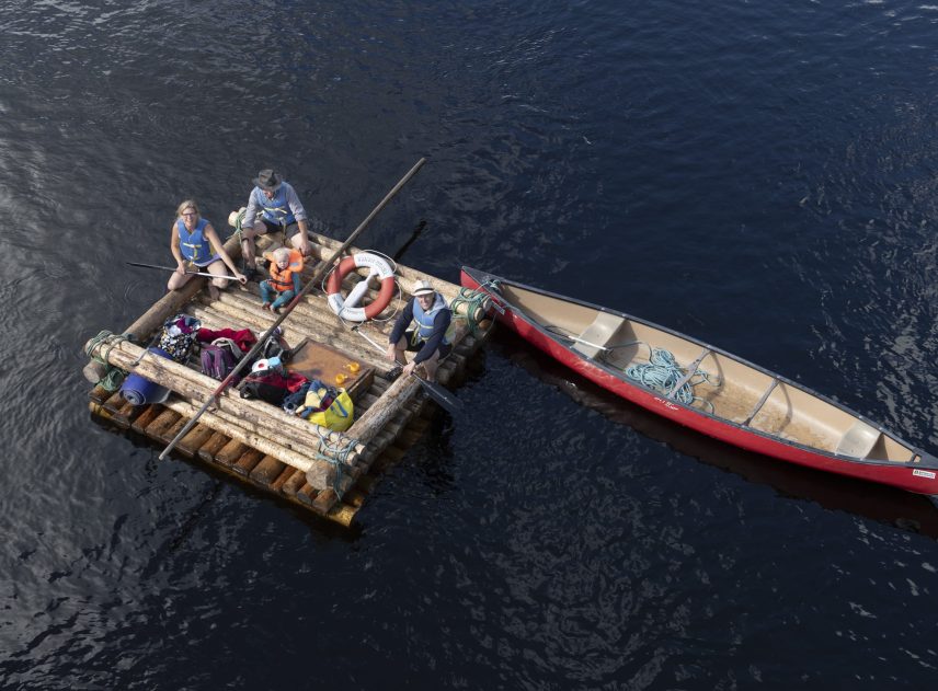 En luftbild av en mindre timmerflotte som flyter på det mörkblå vatten. Förtöjt vid flotten är en röd kanot som är längre än flotten. I kanoten ligger några rep.