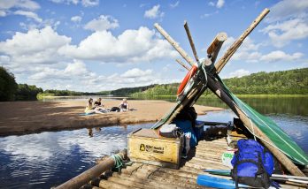 En fullpackad timmerflotte på Klarälven nära en sandbank. På sandbanken sitter tre kvinnor för fika. En strålande sommardag med moln på himlen.