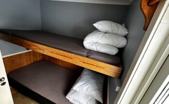 I ett litet rum syns en våningssäng med ordentligt bäddade sängar. Täcken och kuddar ligger i en hög på sängarnas fotända. Till höger skyms ett fönster.