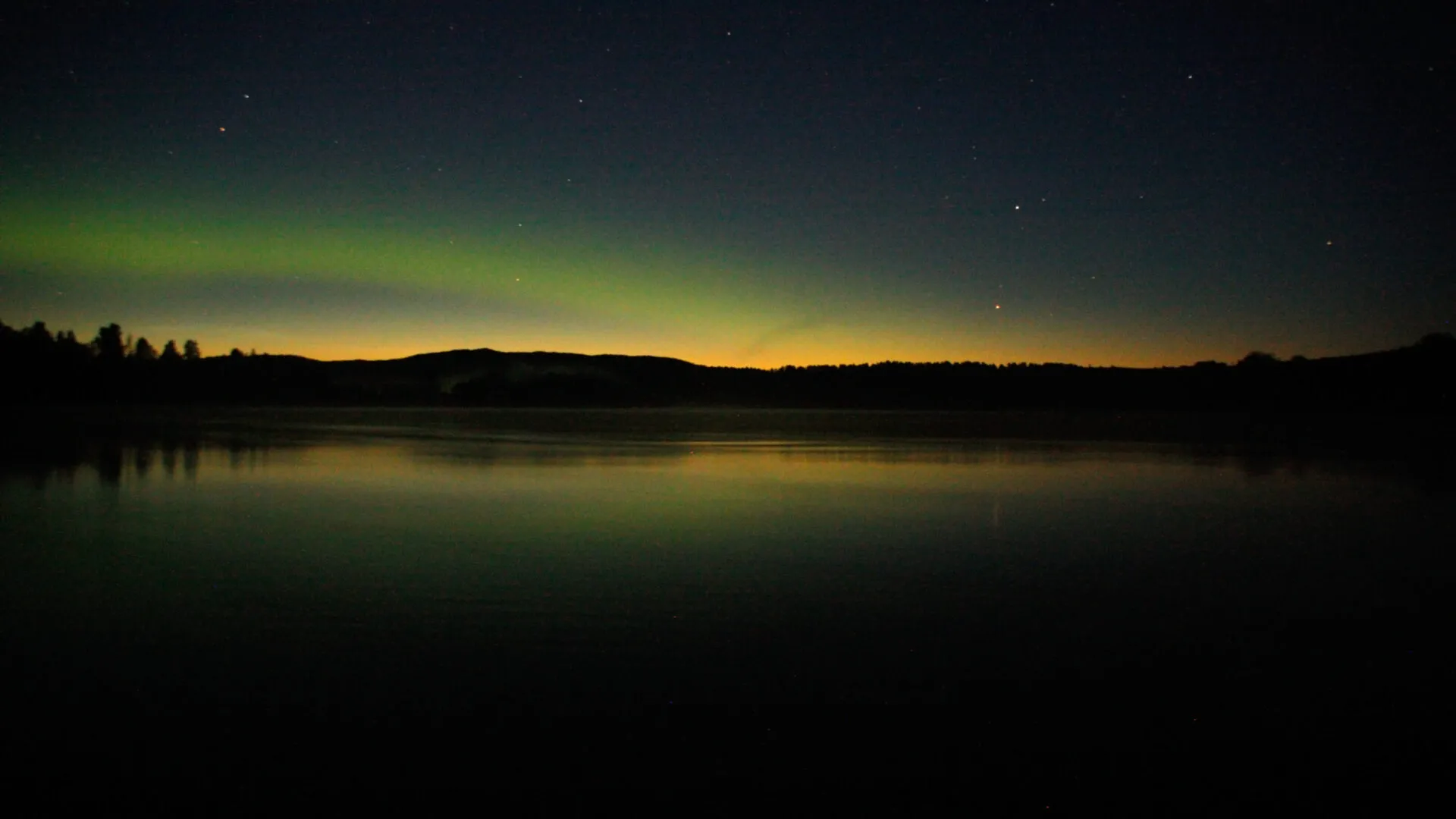 Natten har lagt sig över Klarälven. Bakom träden skyms en sista strimma ljus. En del av himlen är upplyst av ett norrsken, i övrigt blinkar några stjärnor. Skogen syns bara som en siluett.