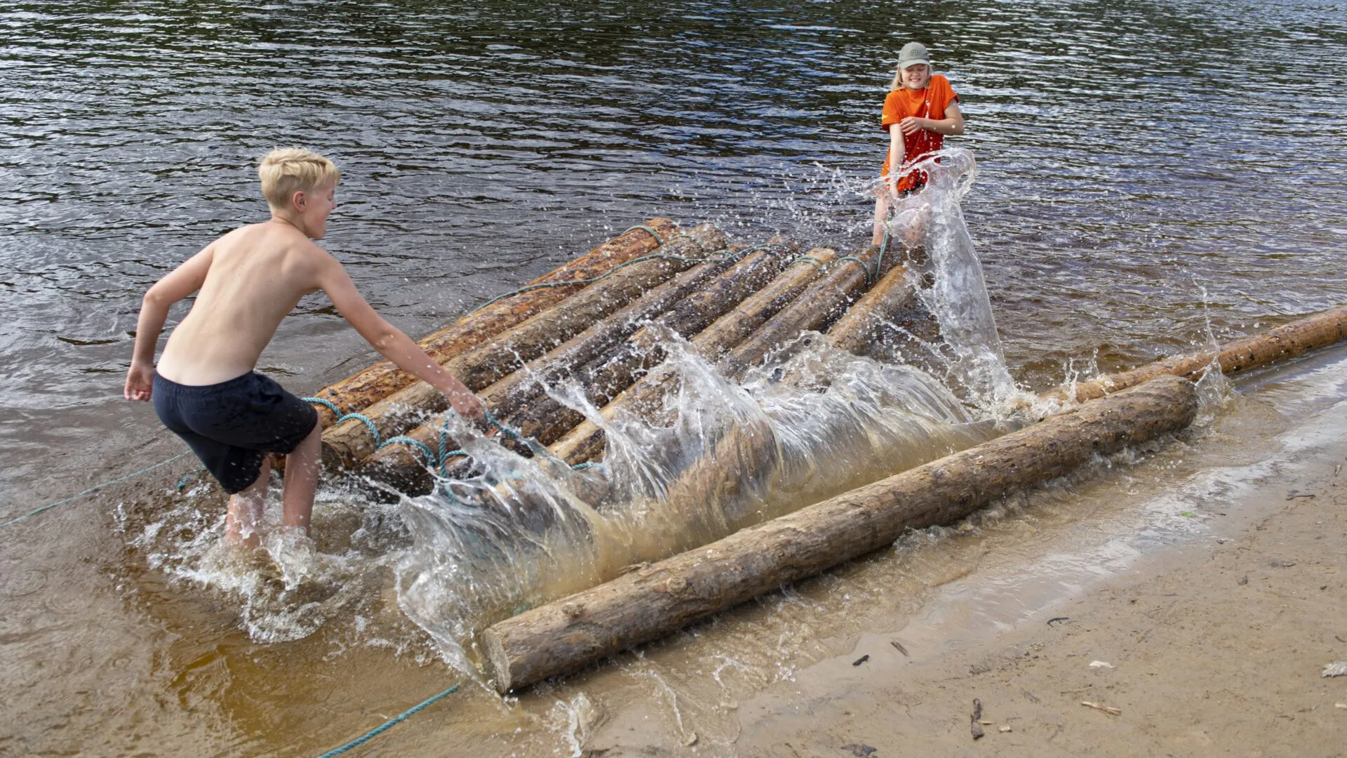 Två tonåringar bygger en timmerflotte i Klarälven vid Klarälvens Camping. Vattnet stänker när stockarna rullar ner från stranden. Flottens understa lager är till hälften färdig.