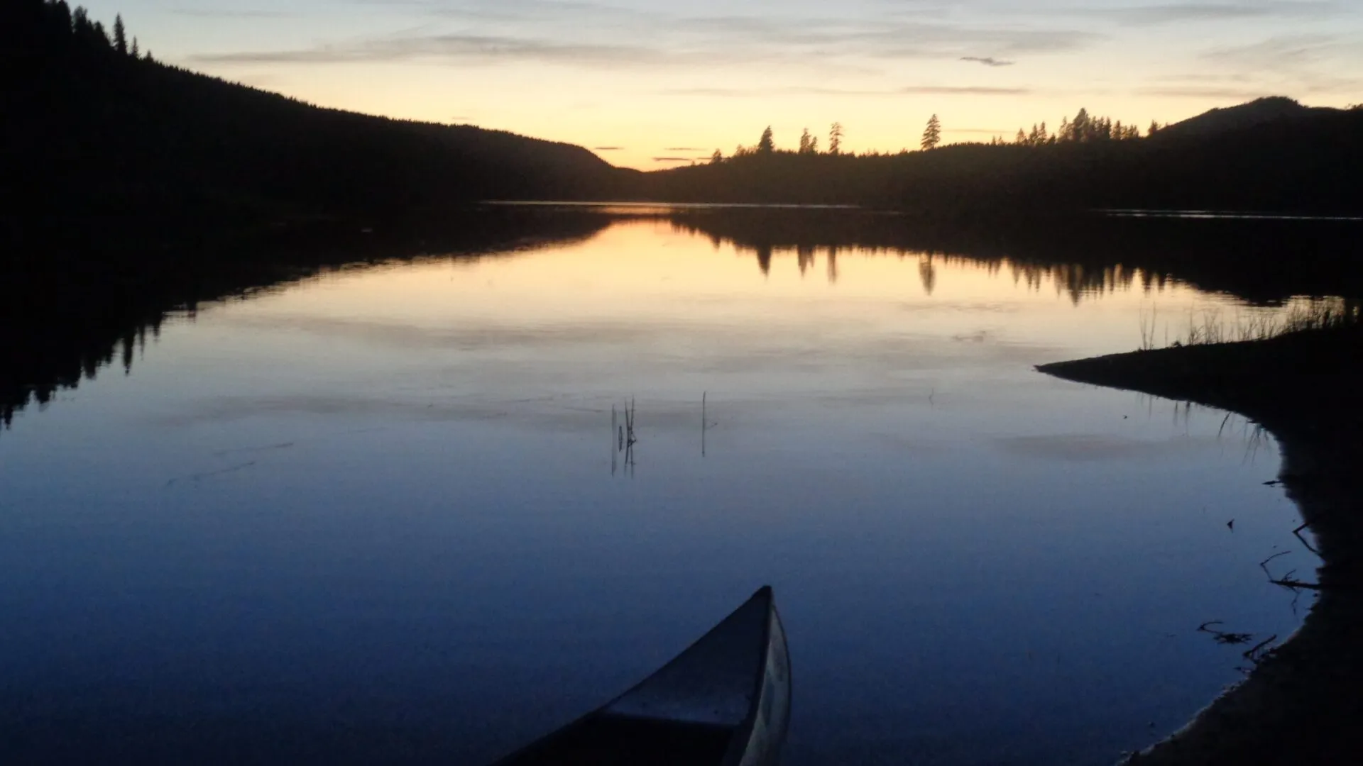 En kanot ligger vid stranden i norra Värmland. Vid horisonten skymmer kvällen, vattnet ligger som en spegel.