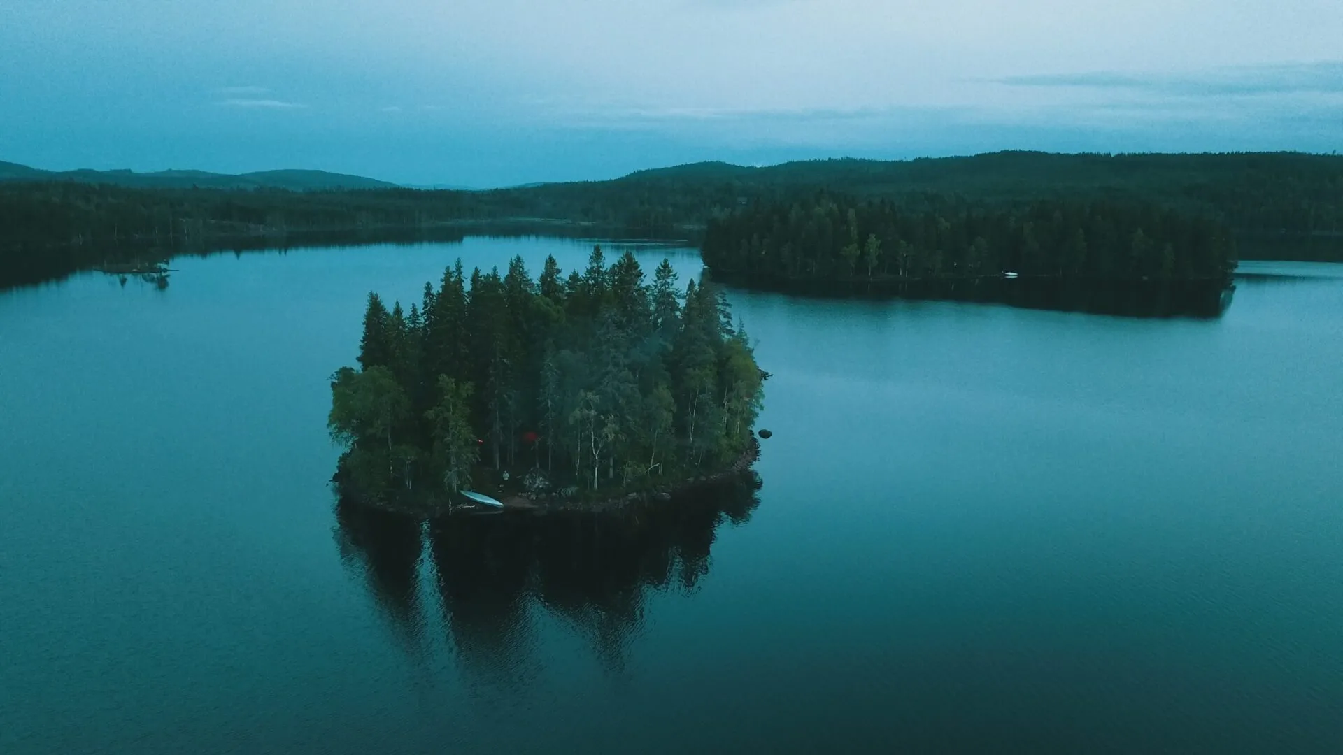 En stor sjö som hör till sjösystemet i Bergslagen breder ut sig med turkosblått vatten. En ö ligger mitt i sjön. I en glänta på ön syns en kanot. Landskapet bakom sjön är klätt i en mörkgrön skog. 