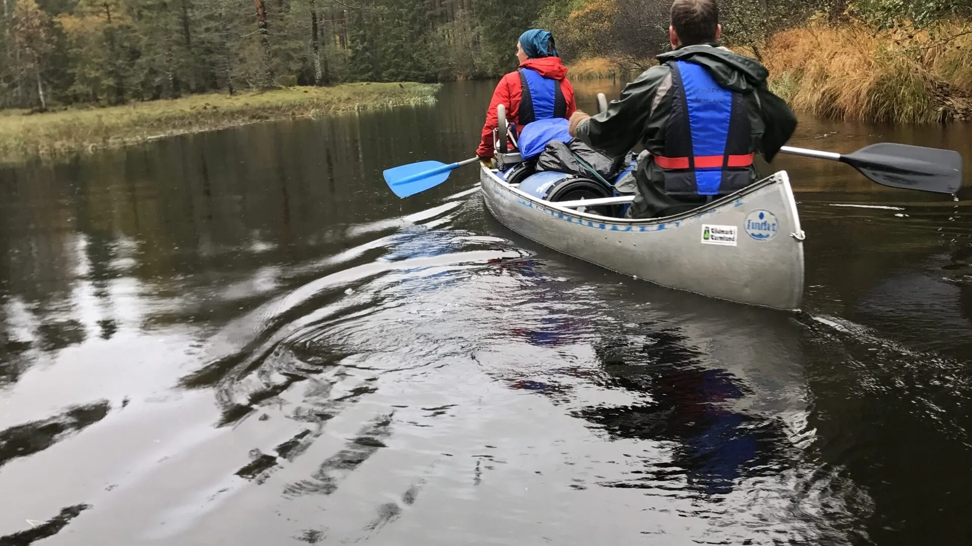 Två personer paddlar på Svartälven i östra Värmland. Kanoten är lastad med mycket packning. Det verkar vara en regnig dag, trädens löv avslöjar en annalkande höst. 