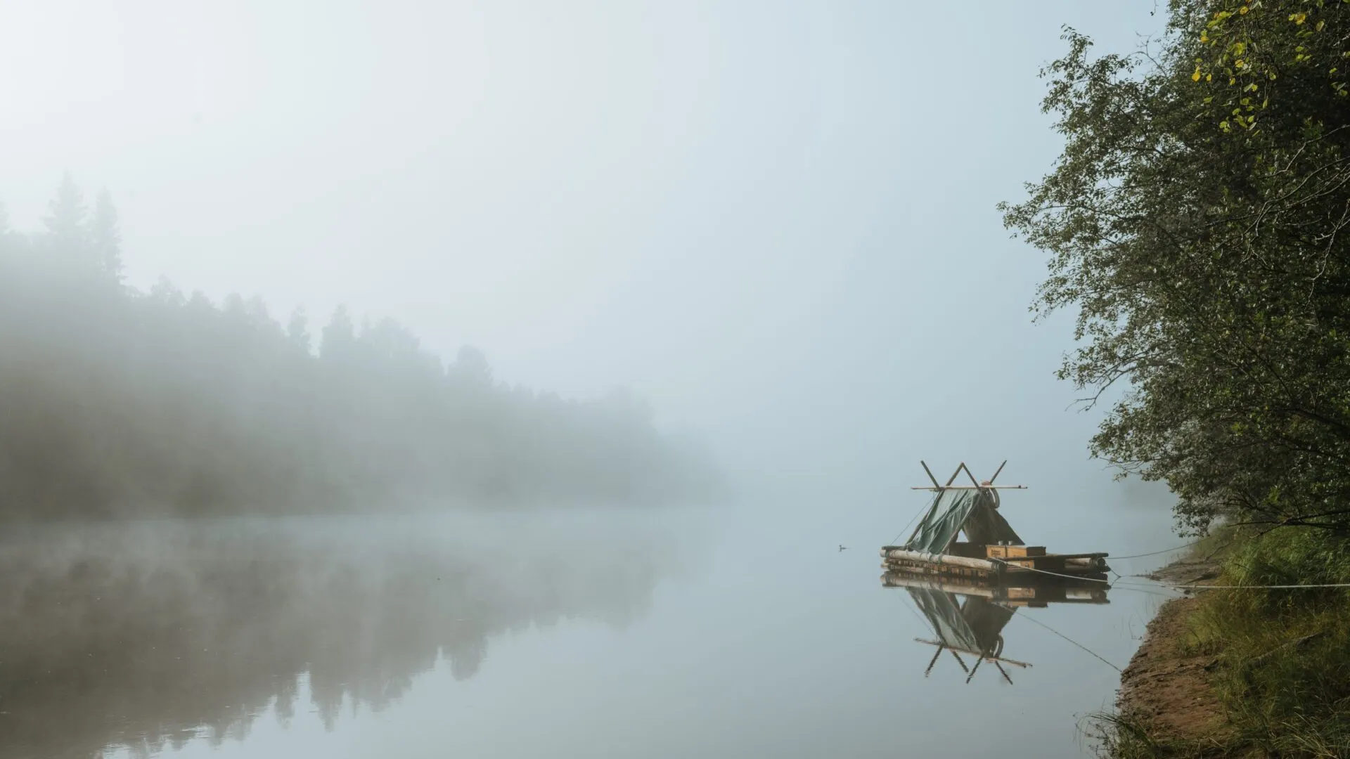 Der Klarälven liegt in starkem Nebel, Himmel und Wasser haben die gleiche Farbe. Ein Holzfloß mit Zelt ist am rechten Ufer vertäut. Das gegenüberliegende Ufer ist nur zu erahnen.
