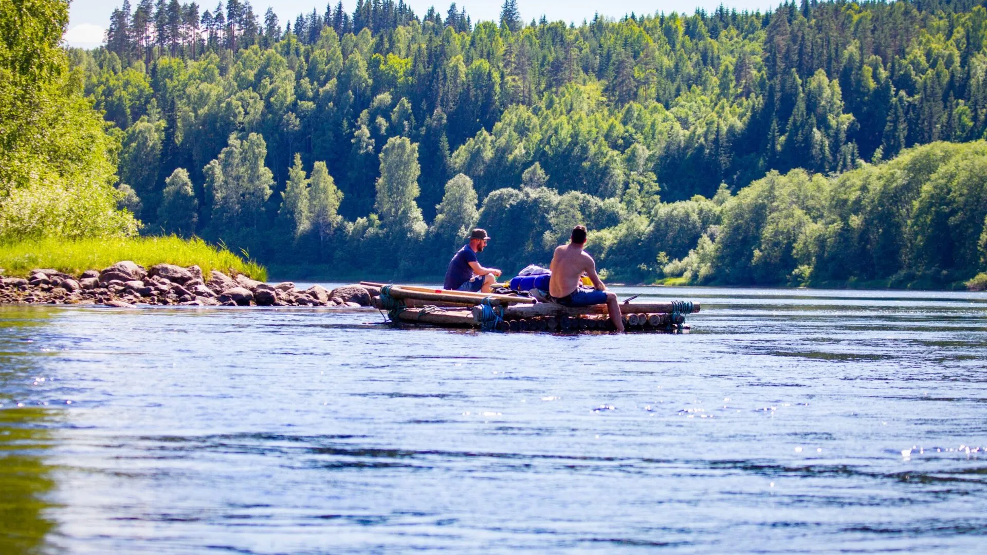 Två män sitter ombord på en mindre timmerflotte utan tältkonstruktion. En varm solig sommardag. Flotte glider lugnt och fint nedströms, männen doppar fötterna i vattnet. Mycket grönska längs strandkanten.